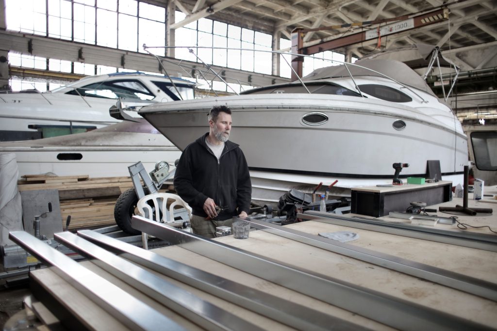 Concentrated bearded adult mechanic in casual clothes working with metal details near boat in garage